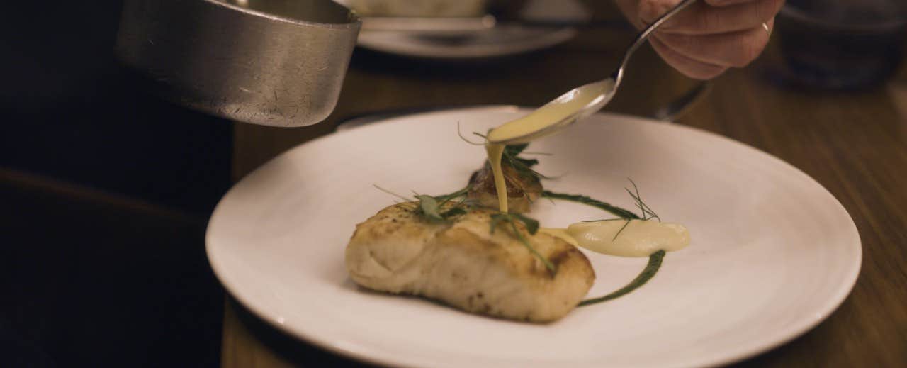 A chef pouring sauce over a piece of grilled fish garnished with herbs on a white plate.