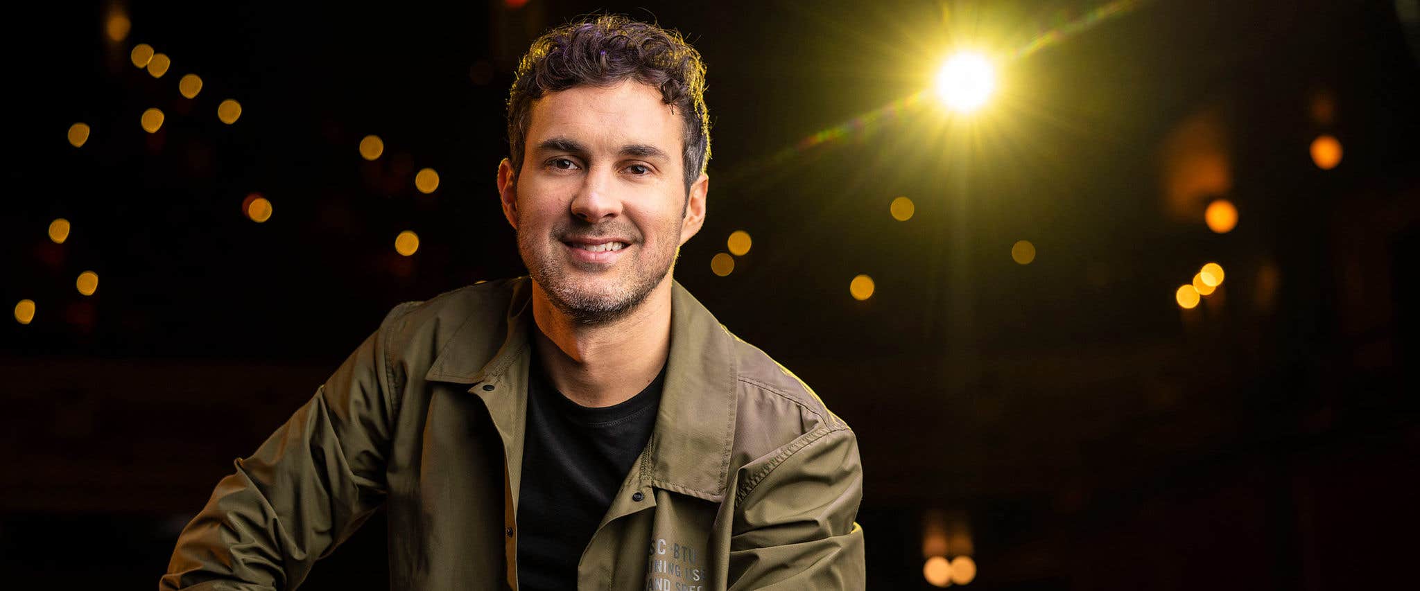 Mark Normand in a black shirt and olive/brown colored jacket against a black background with some out of focus lights and a bright yellow spotlight