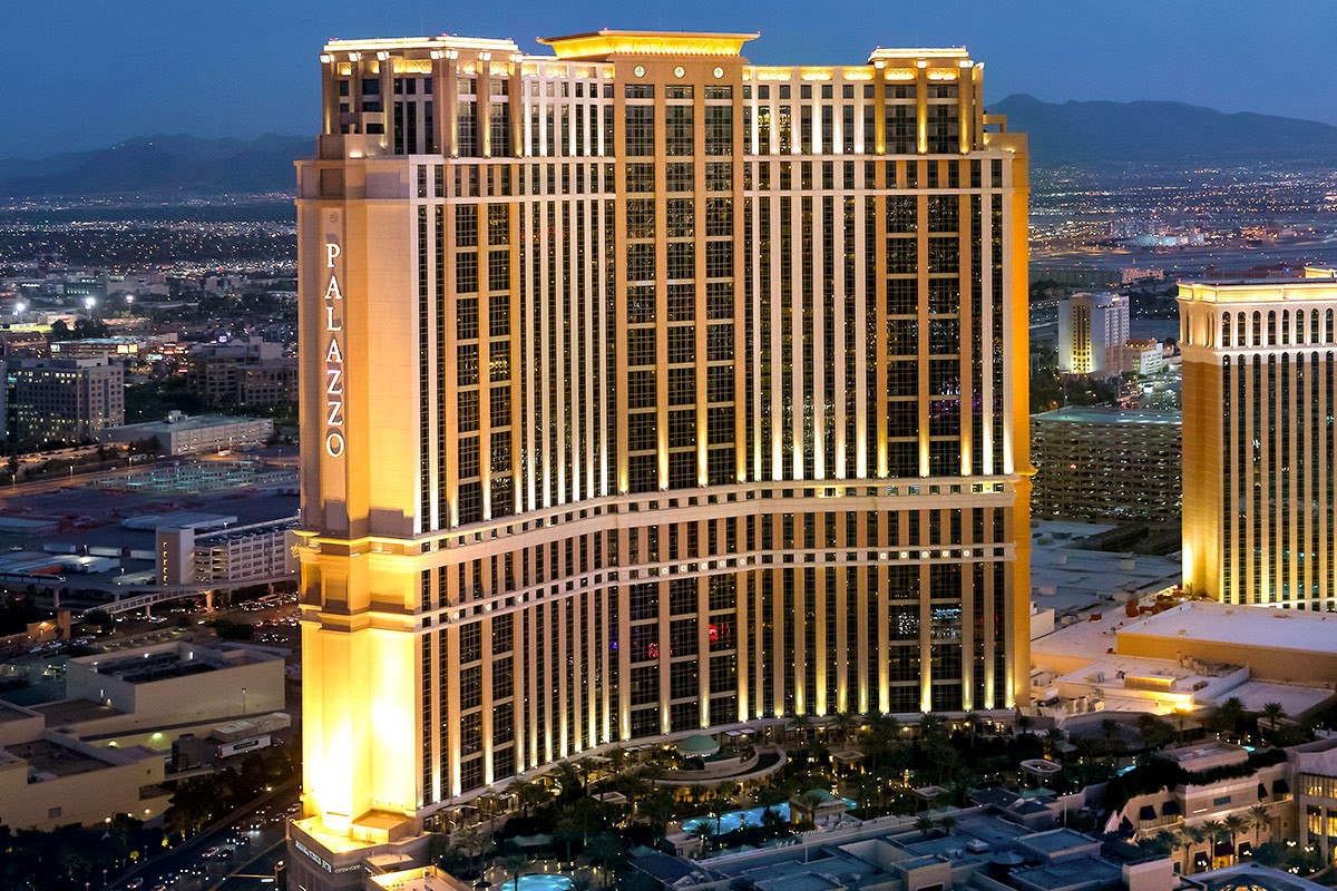 Aerial view of The Palazzo resort in Las Vegas, brightly lit at dusk with surrounding cityscape and distant mountains.