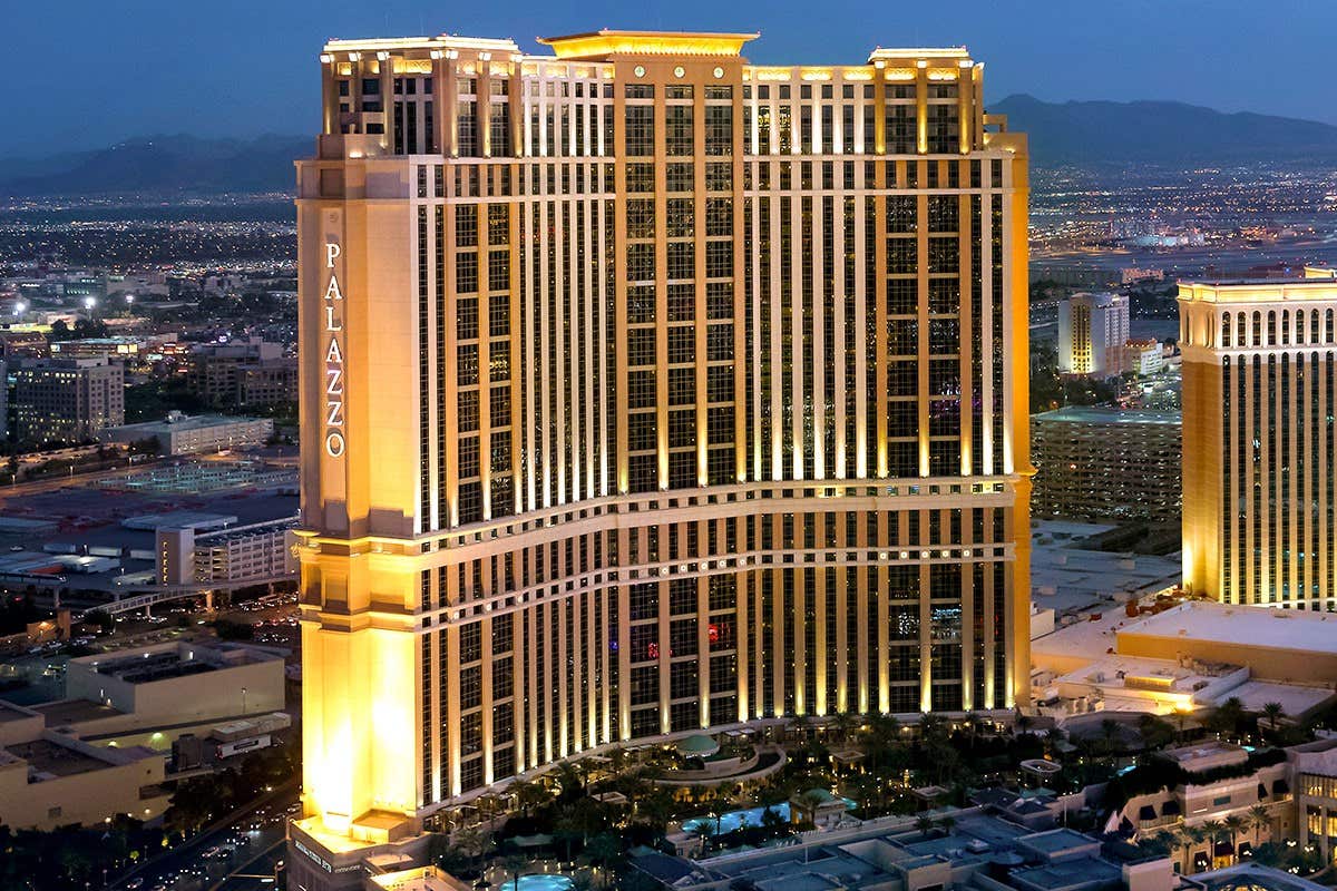 Aerial view of The Palazzo hotel in Las Vegas, brightly lit at dusk with surrounding cityscape and distant mountains.