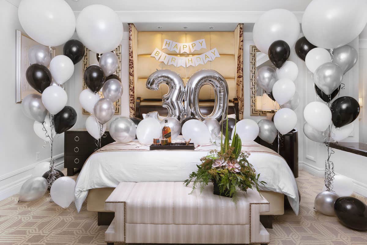 A room decorated for a 30th birthday with balloons and a "Happy Birthday" banner over a bed with a bouquet at the foot.