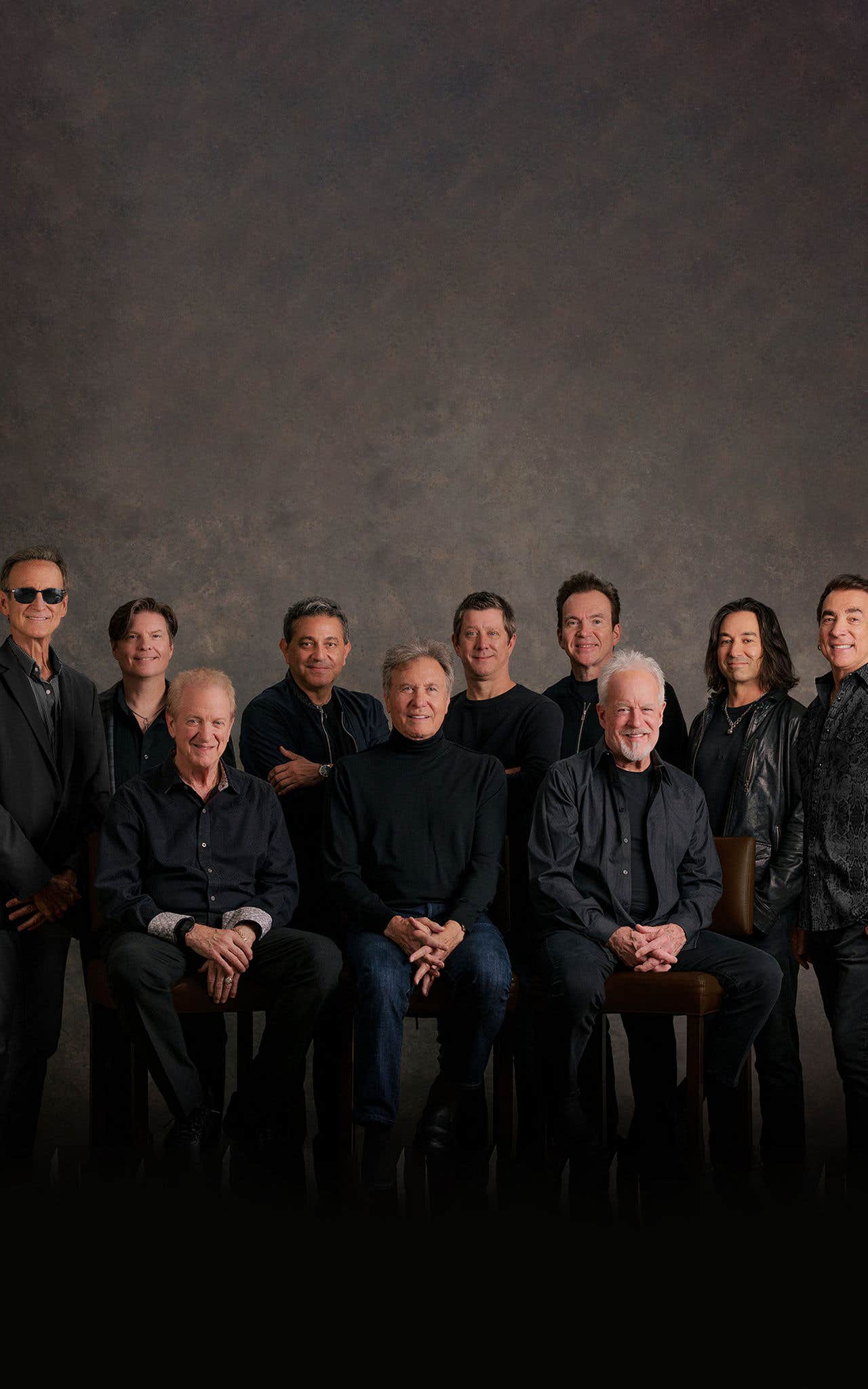 Group photo of Chicago, all dressed in black or dark clothing, standing and seated in front of a dark background.