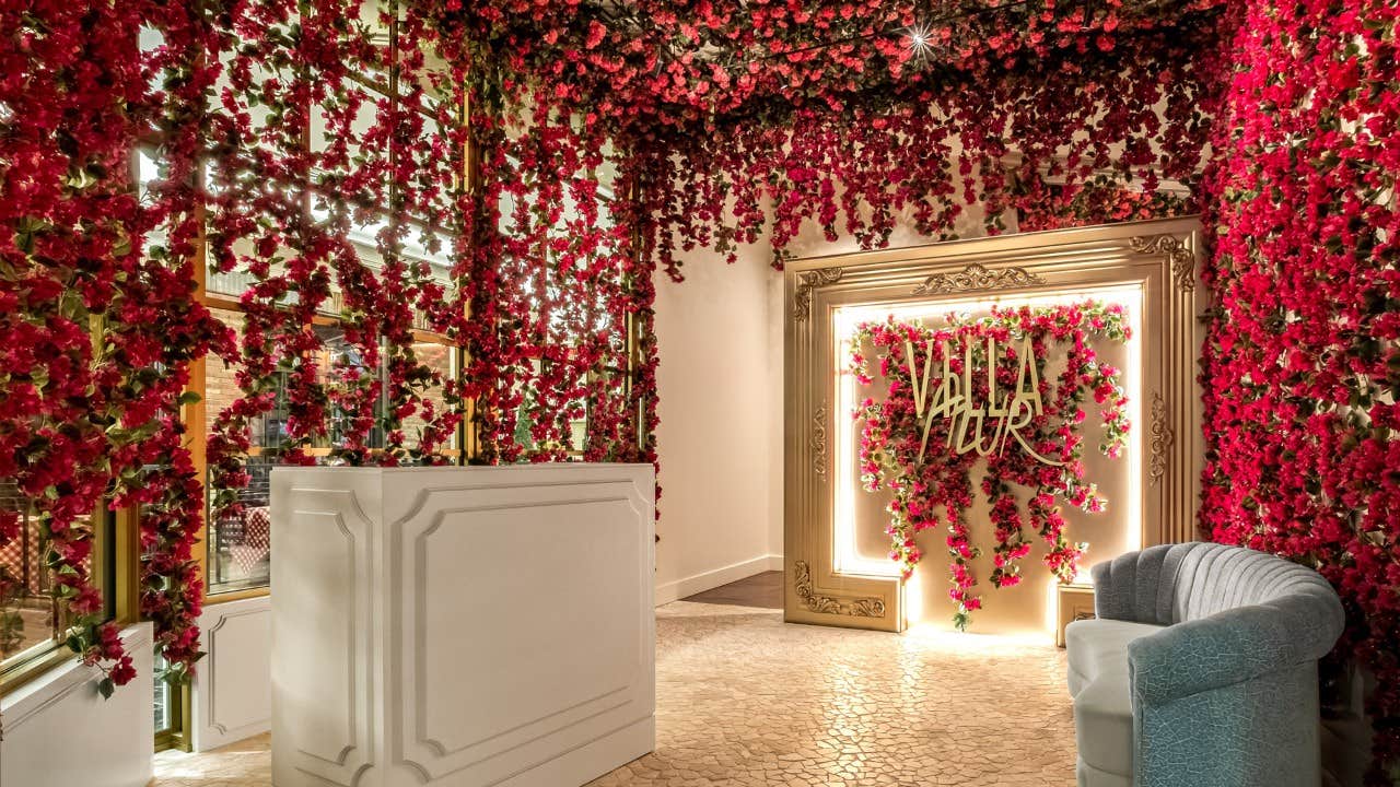 A lobby adorned with vibrant red flowers, a white counter, a grey chair, and a gold-framed mirror with illuminated decor.