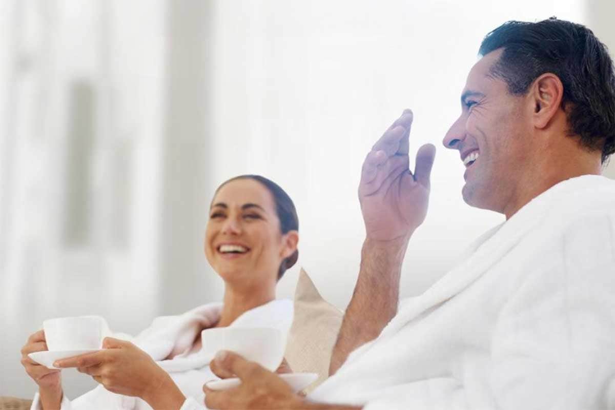 A couple in white robes in a brightly naturally lit room, sharing a laugh with a cup and saucer set in each of their hands