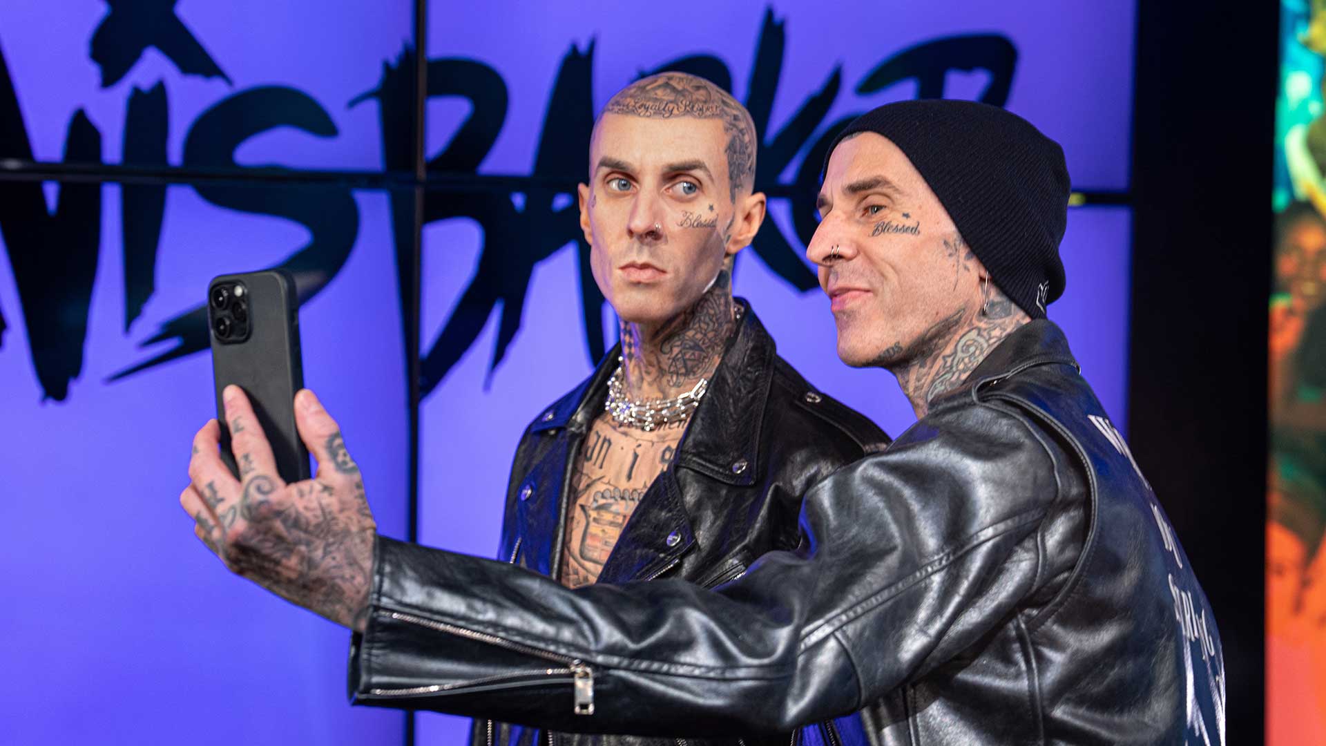 Two tattooed men in leather jackets take a selfie in front of a colorful background with bold text.