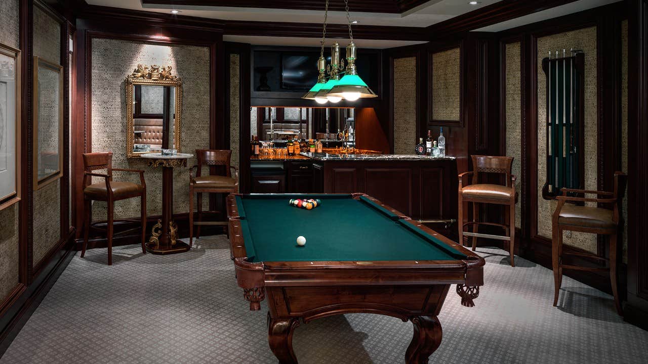 A stylish room with a pool table, bar, bar stools, ornate wall mirror, and green lamp over the pool table.