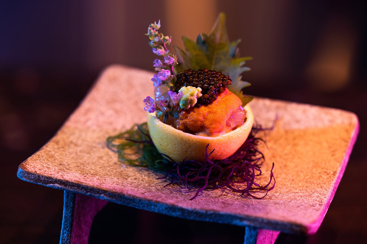 Elegant dish with caviar, herbs, and uni served in a lemon bowl on a textured stone plate.