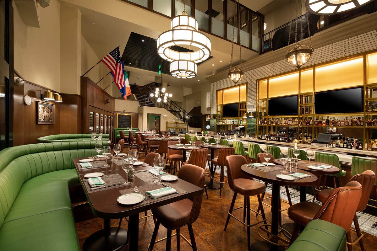 Upscale restaurant with green booths, brown chairs, elegantly set tables, multiple flags, and a bar with warm lighting.