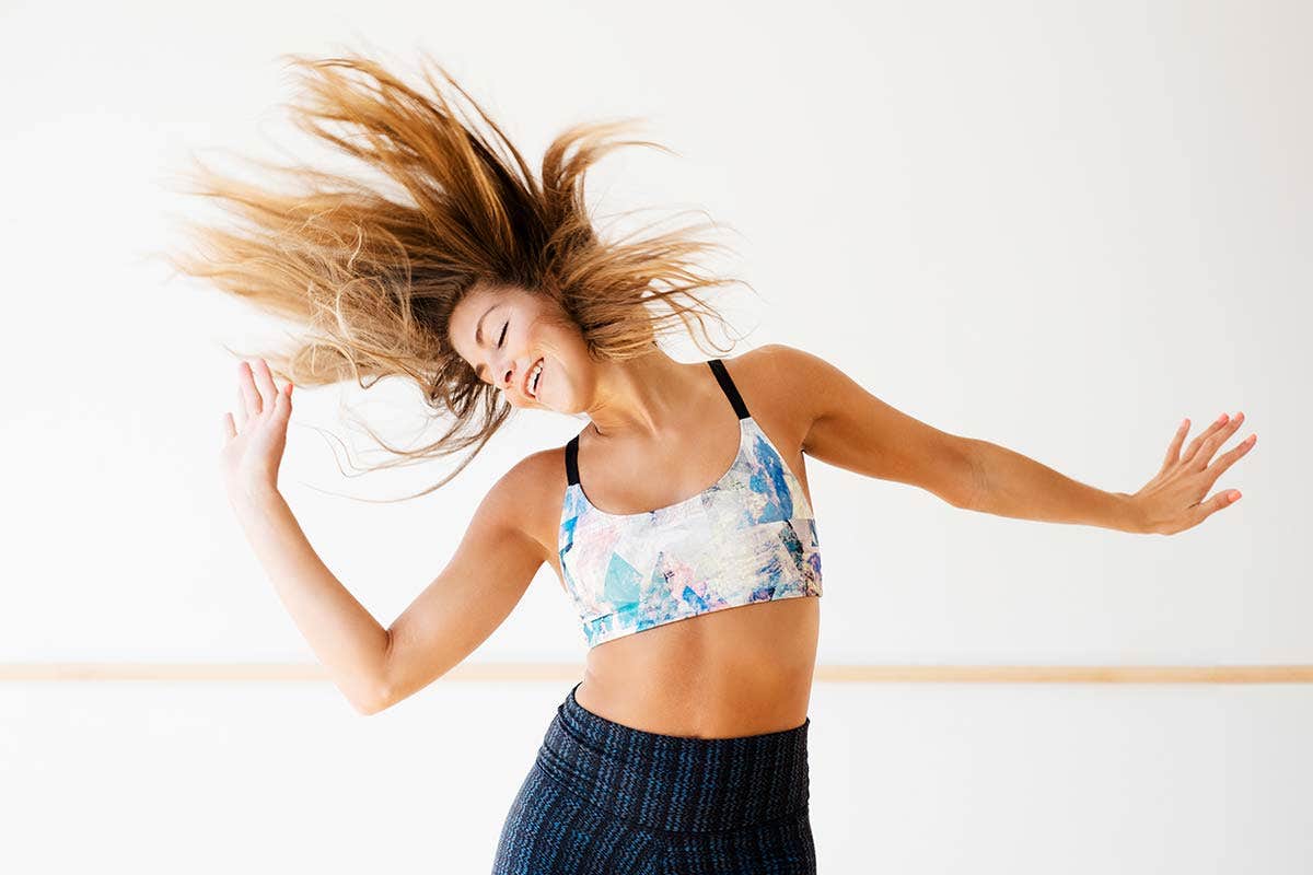 A woman dancing with her hair flipping in the air, wearing a sports bra and leggings in a bright studio.