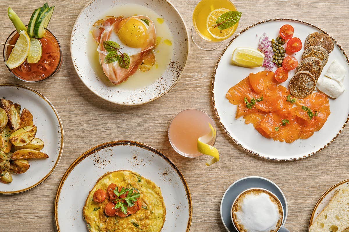 A table with various brunch foods, including eggs Benedict, smoked salmon, roasted potatoes, an omelet, and cocktails.