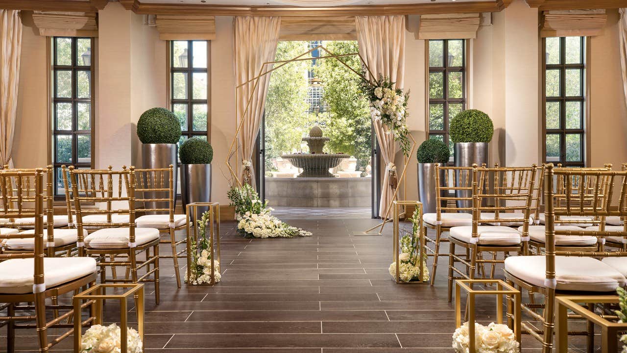 An elegant indoor wedding venue with gold chairs, white floral arrangements, and a view of a fountain outside.