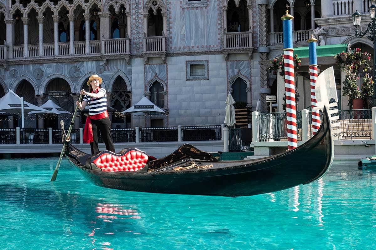 A gondolier rows a gondola with red checkered seats in a blue canal in front of a Venetian-style building.