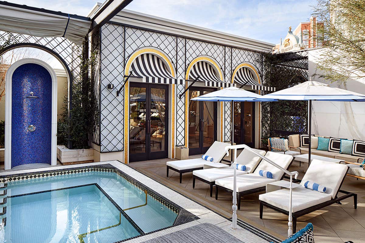 Outdoor patio with a small pool, striped sun loungers, umbrellas, blue-tiled fountain, and a chic building with arched windows.