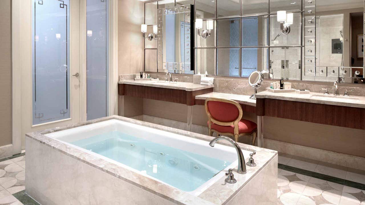 Luxurious bathroom with a large bathtub, double sinks, a vanity area with a red chair, and elegant mirrors on the wall.