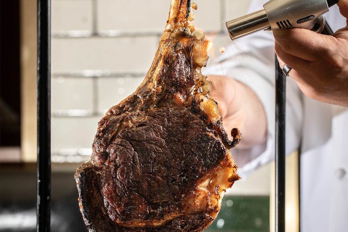 Close-up of a chef holding a large cooked tomahawk steak while using a blow torch to sear it.