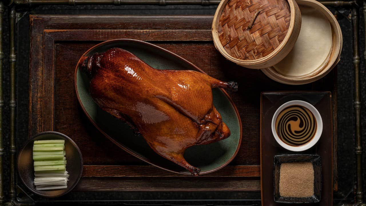 Whole roasted duck with dipping sauce, green onions, pancakes, and brown sugar on a wooden tray with bamboo container.