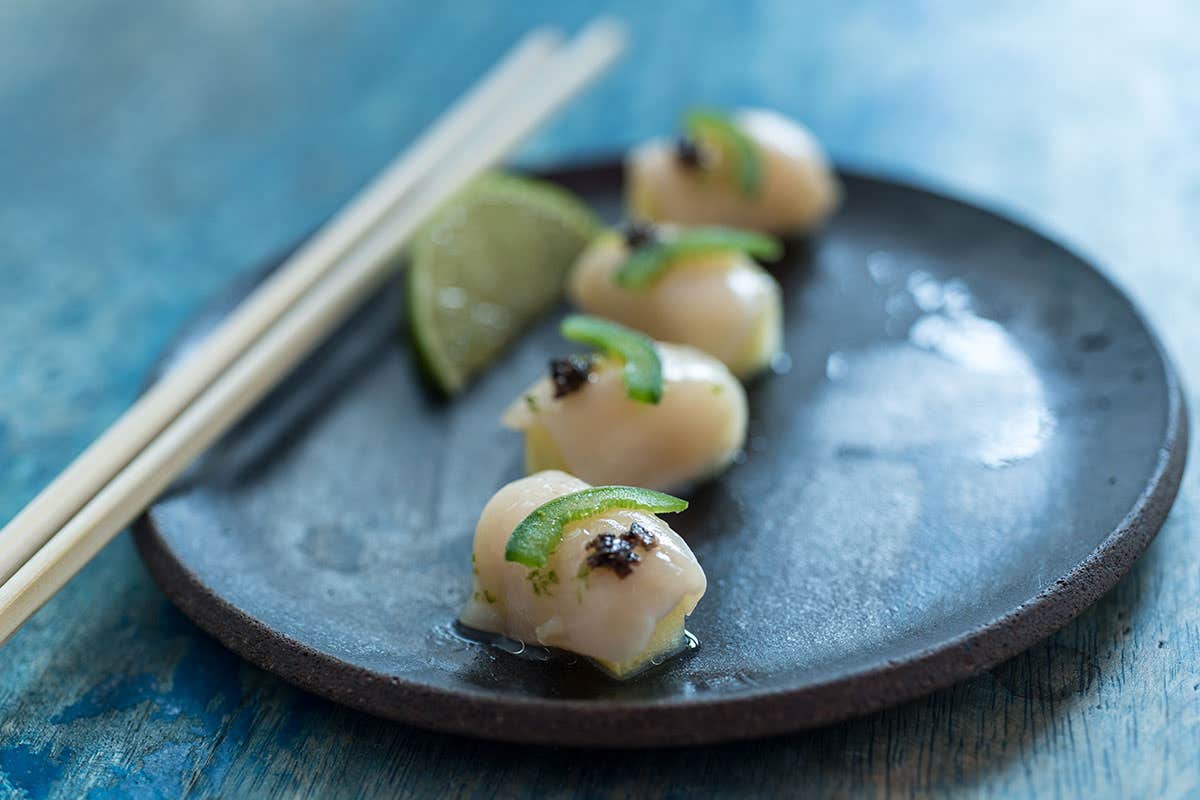 Four scallops garnished with sliced green pepper and black seasoning on a black plate with chopsticks and lime wedges.