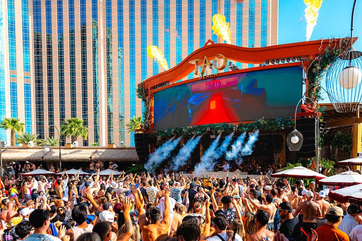 A crowd in front of the stage at TAO Beach, which is blowing fog machines downward, and above the stage, there are flames blasting out upwards