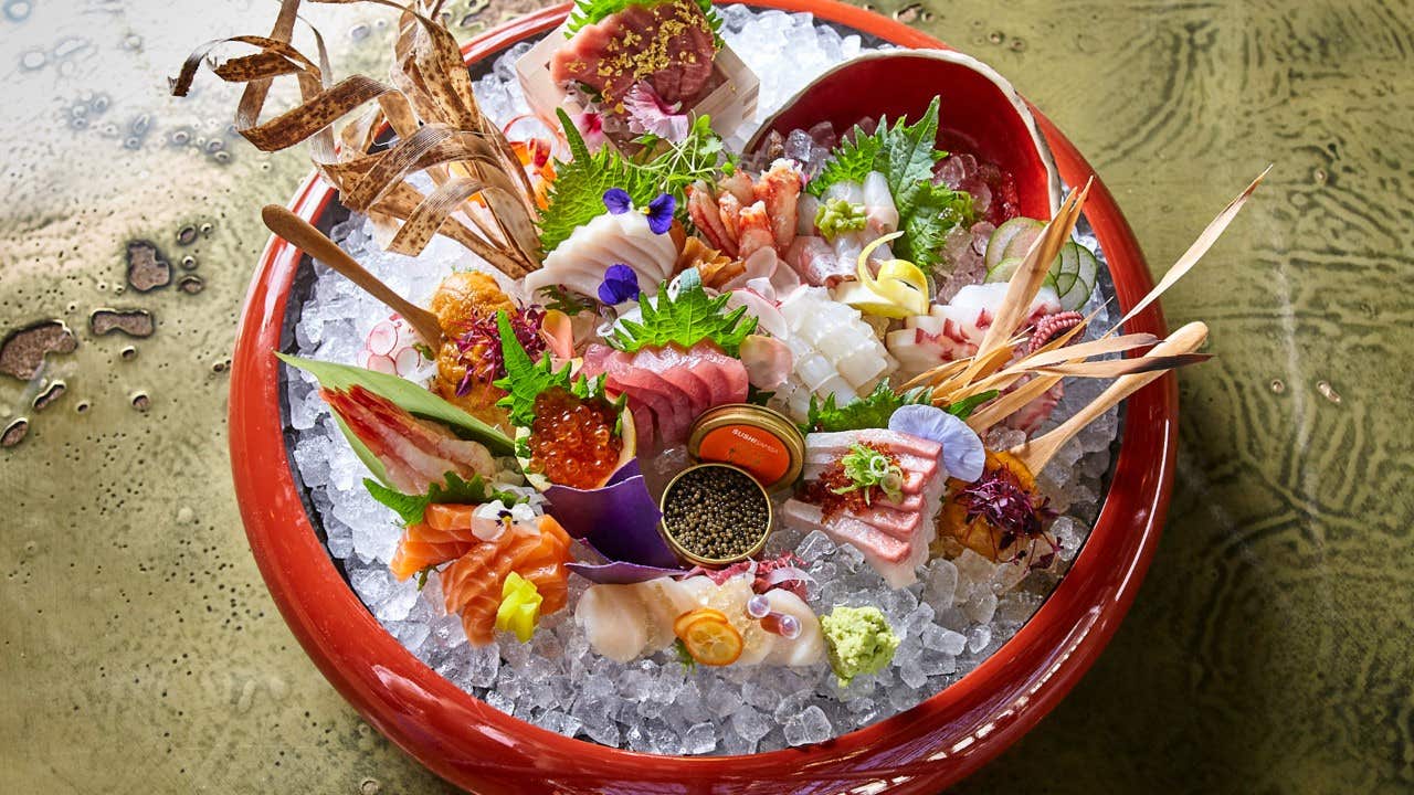 A vibrant sashimi platter with assorted seafood, garnishes, and flowers on ice in a red bowl.