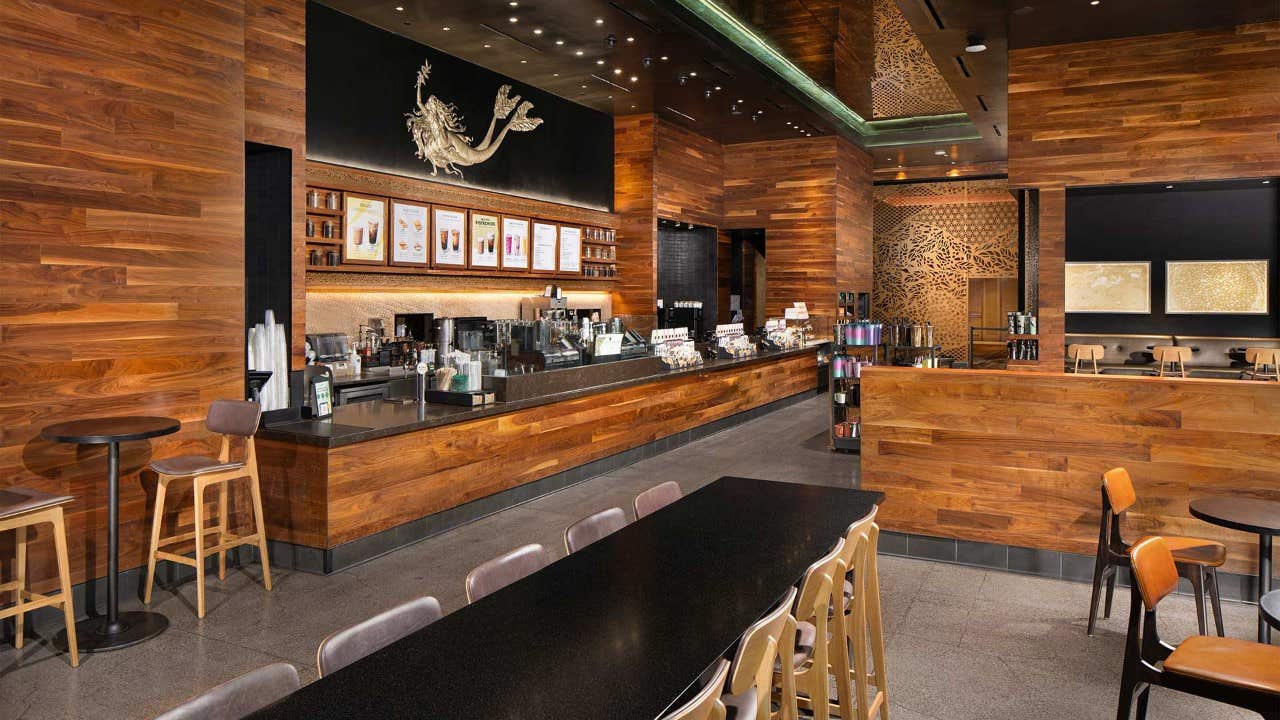 Interior of a modern coffee shop with wooden walls, a long black table, bar stools, and a well-lit counter area.