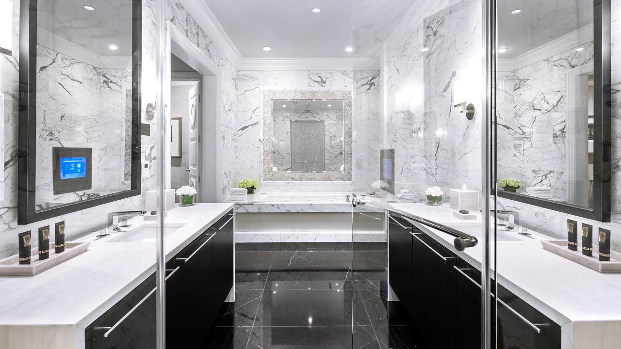 Luxurious modern bathroom with marble walls and floor, double sinks, a soaking tub, large mirrors, and sleek black cabinets.