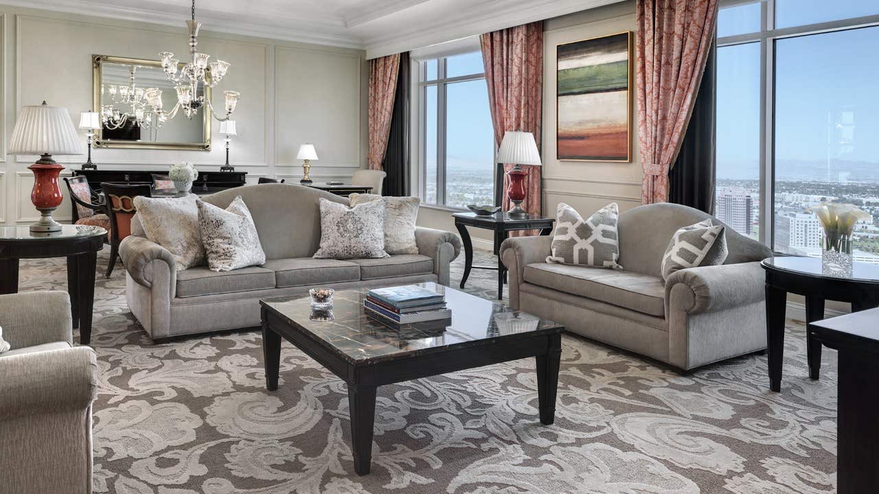 Luxurious living room with elegant furniture, chandeliers, large windows with drapes, and a cityscape view in the background.