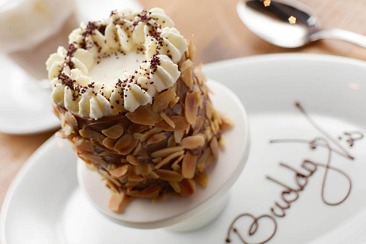 Close-up of a dessert with almond slices and whipped cream on a plate with "Buddy V's" written in chocolate.