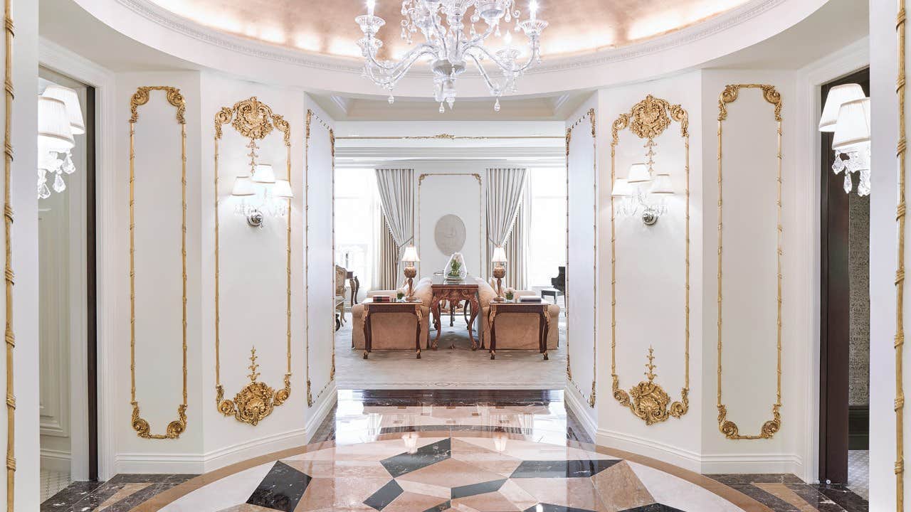 Elegant, ornate hallway with gold details leading to a luxurious sitting area with chandeliers and beige furniture.