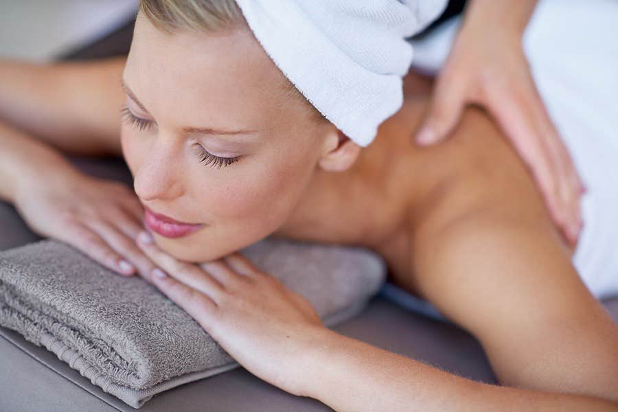 Woman with a towel on her head receiving a back massage, lying on a gray towel with eyes closed, looking relaxed.