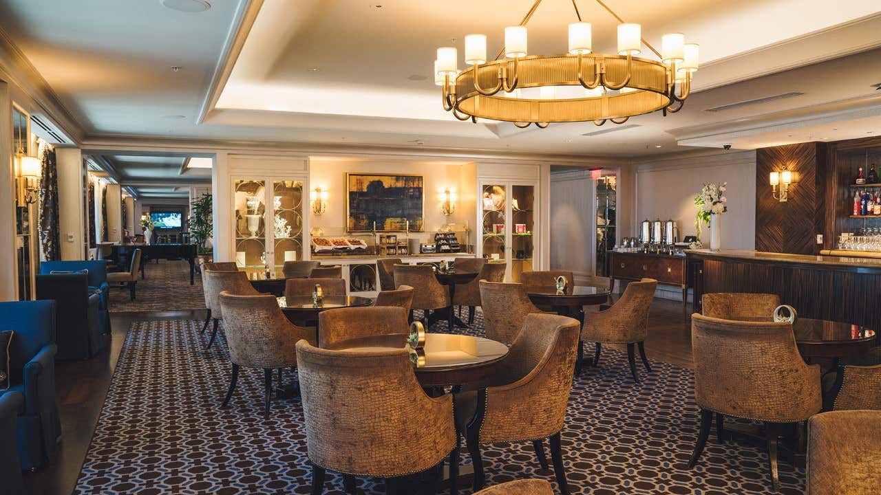 Elegant hotel lounge with circular tables, brown chairs, a chandelier, and a bar area on the right side.