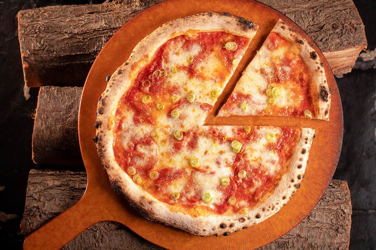 A close-up of a sliced pizza with melted cheese and toppings on a wooden board, placed over stacked firewood.