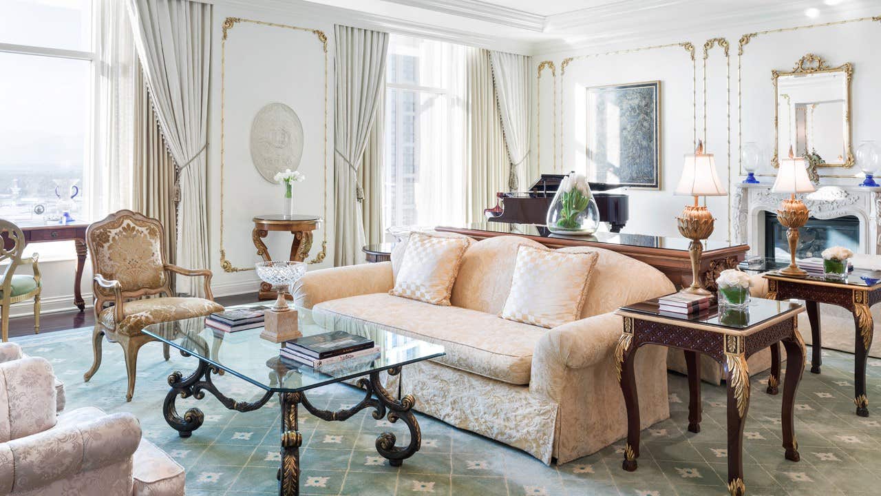 Elegant living room with ornate furniture, large windows with drapes, glass coffee table, and a grand piano by the window.