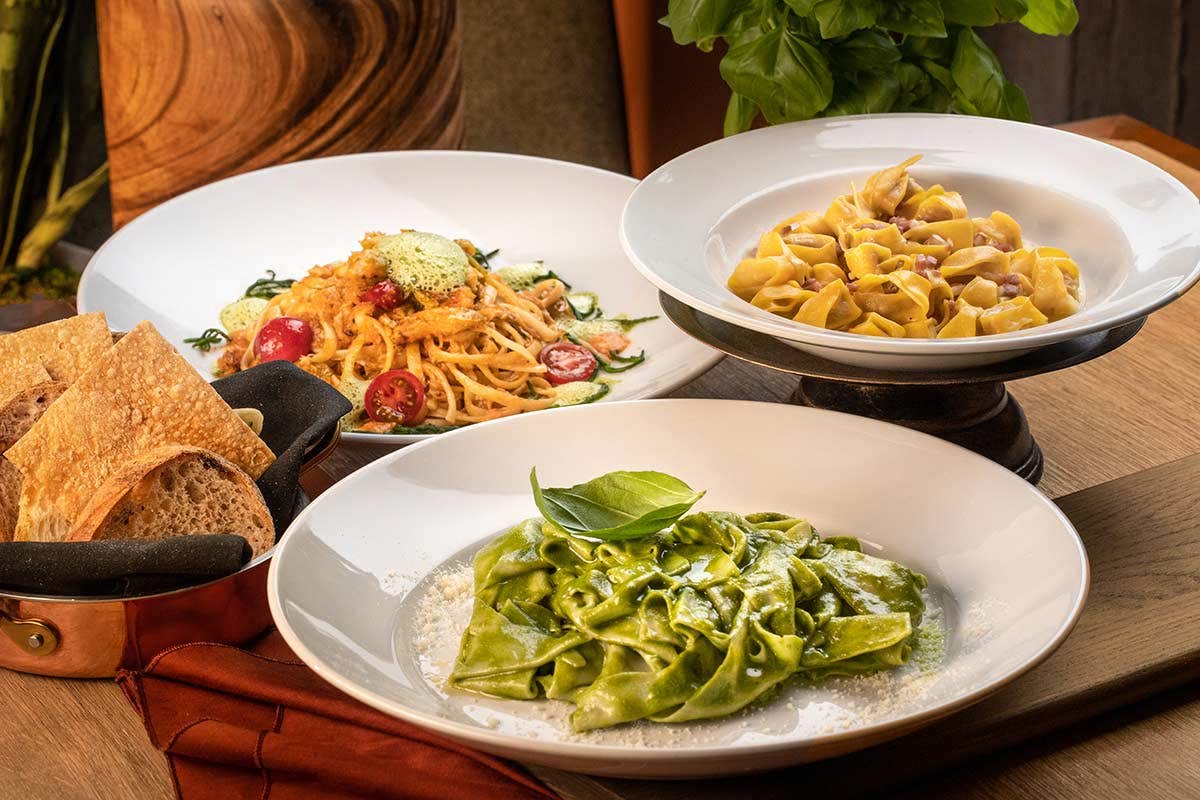 Three dishes of pasta: pesto with basil, tortellini with cream sauce, and spaghetti with tomatoes, next to sliced bread.