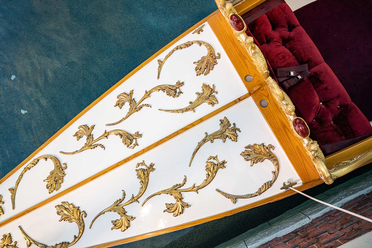 Ornate gondola with gold leaf designs on white panels and red velvet seating, floating on a greenish-blue canal.