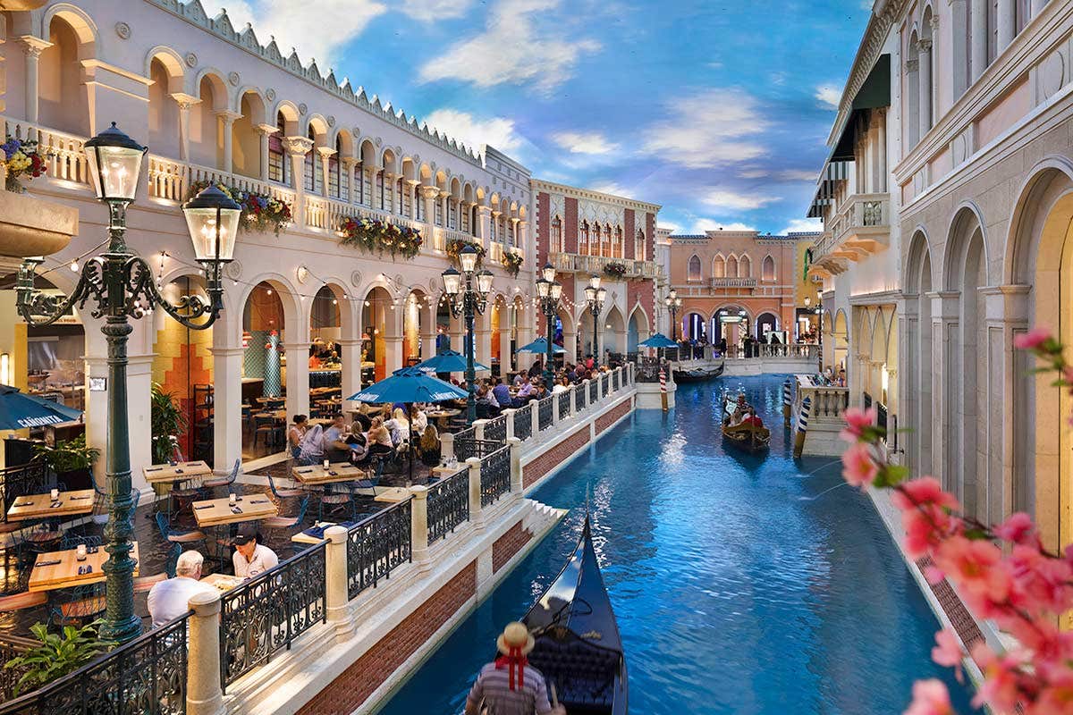 People dine by a canal with gondolas in an indoor setting resembling Venice, under a painted blue sky.