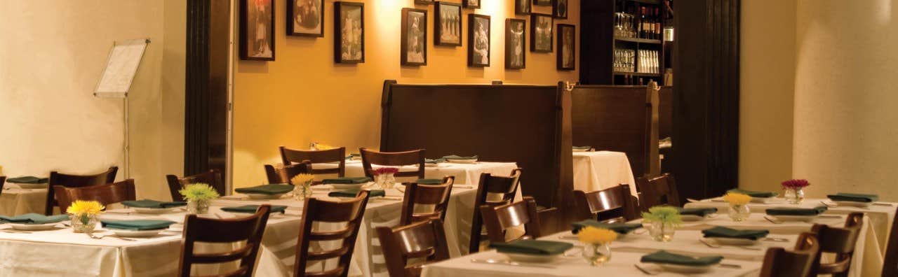 Cozy restaurant with yellow walls, wooden chairs, white tablecloths, and framed photos on the wall.