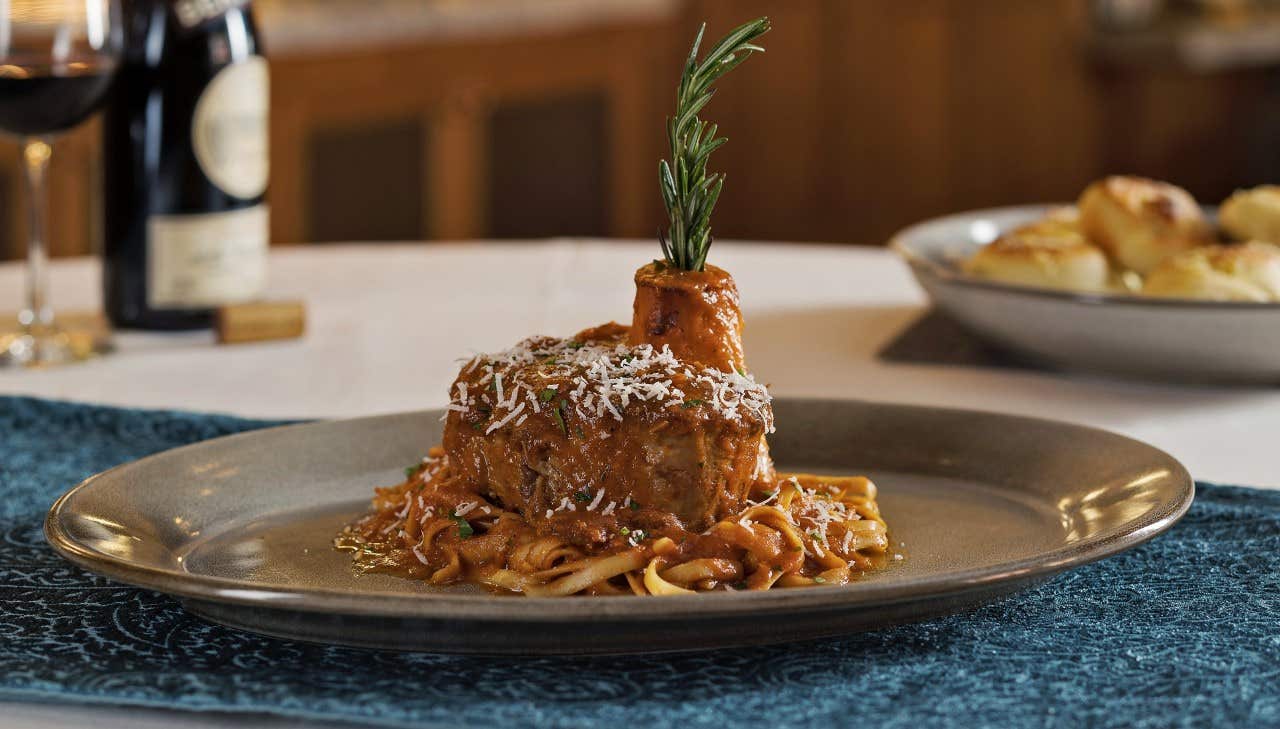 A plated dish of osso buco garnished with rosemary, served over pasta with grated cheese, with bread and wine in the background.