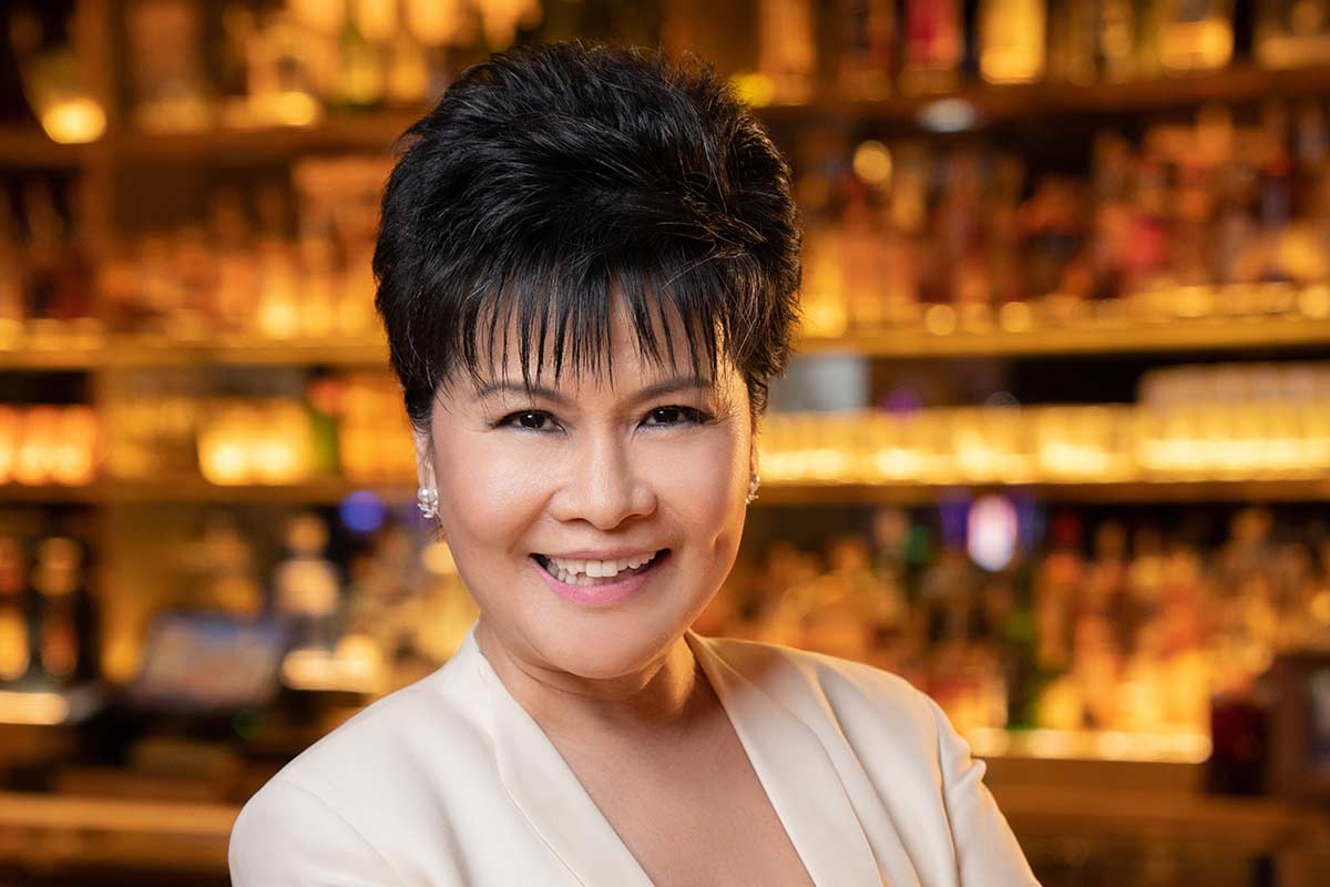 A smiling woman with short black hair wearing a white blazer, standing in front of a bar with bottles in the background.