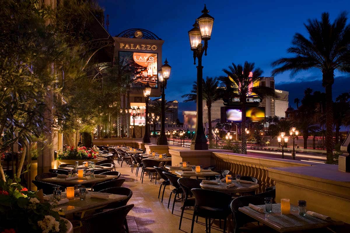 Outdoor restaurant with tables and chairs, illuminated lanterns, palm trees, and city lights at night. Buildings in the background.