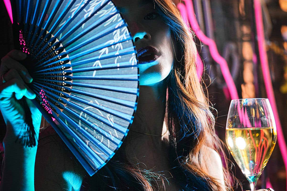 Person holding a blue fan and a glass of wine in a colorful, neon-lit setting.