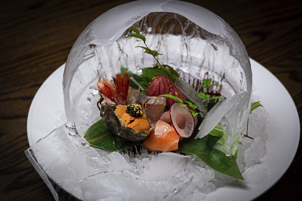 Assorted sashimi beautifully arranged on leaves, surrounded by a dome of ice, served on a white plate.