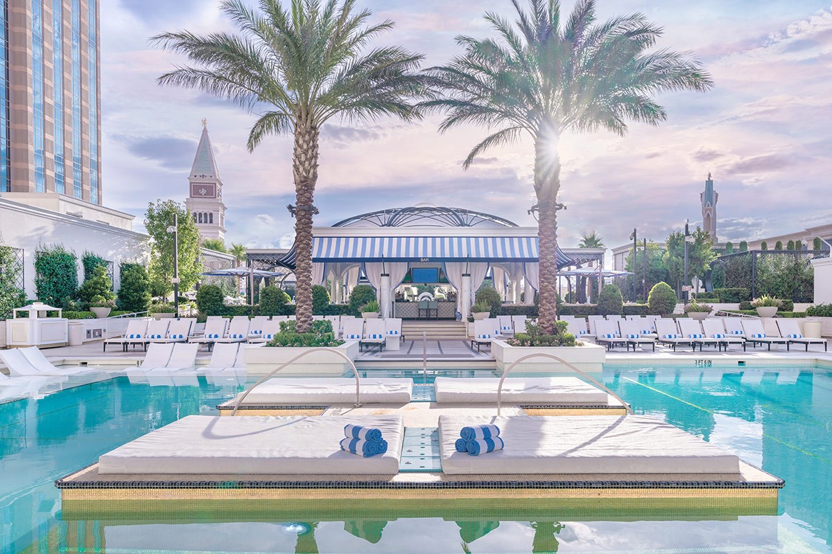 A luxurious poolside area with lounge chairs, cabanas, and palm trees under a pastel sky.