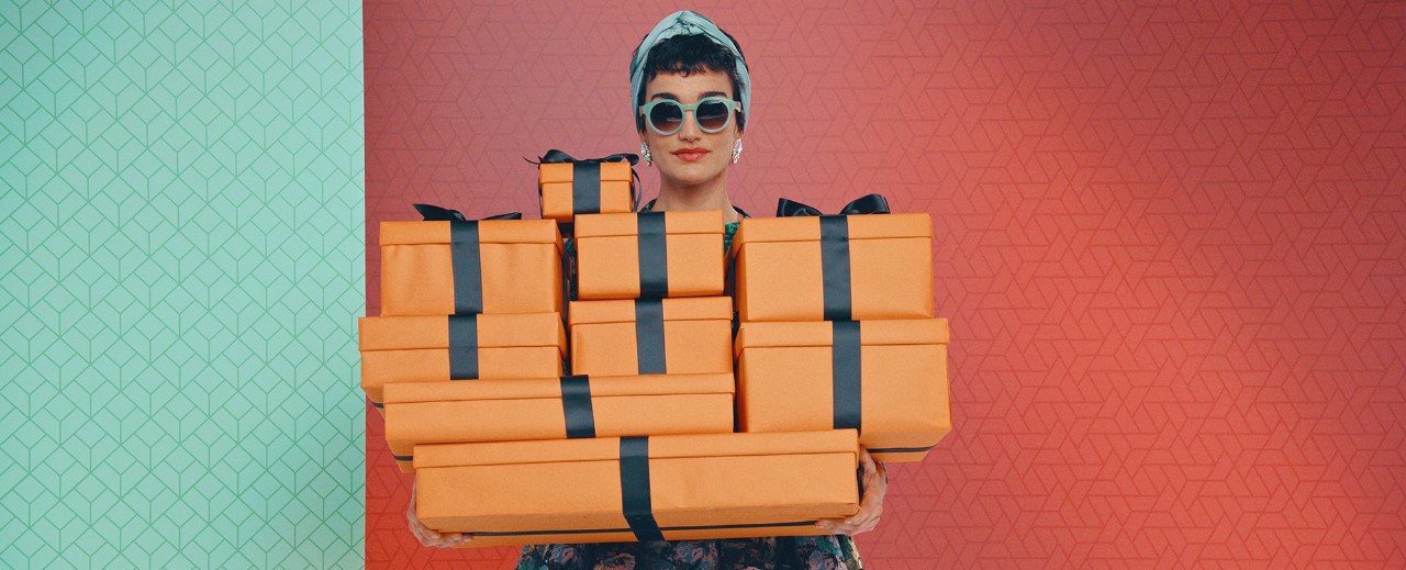 Woman in sunglasses and headscarf holding multiple orange gift boxes with black ribbons against patterned background.