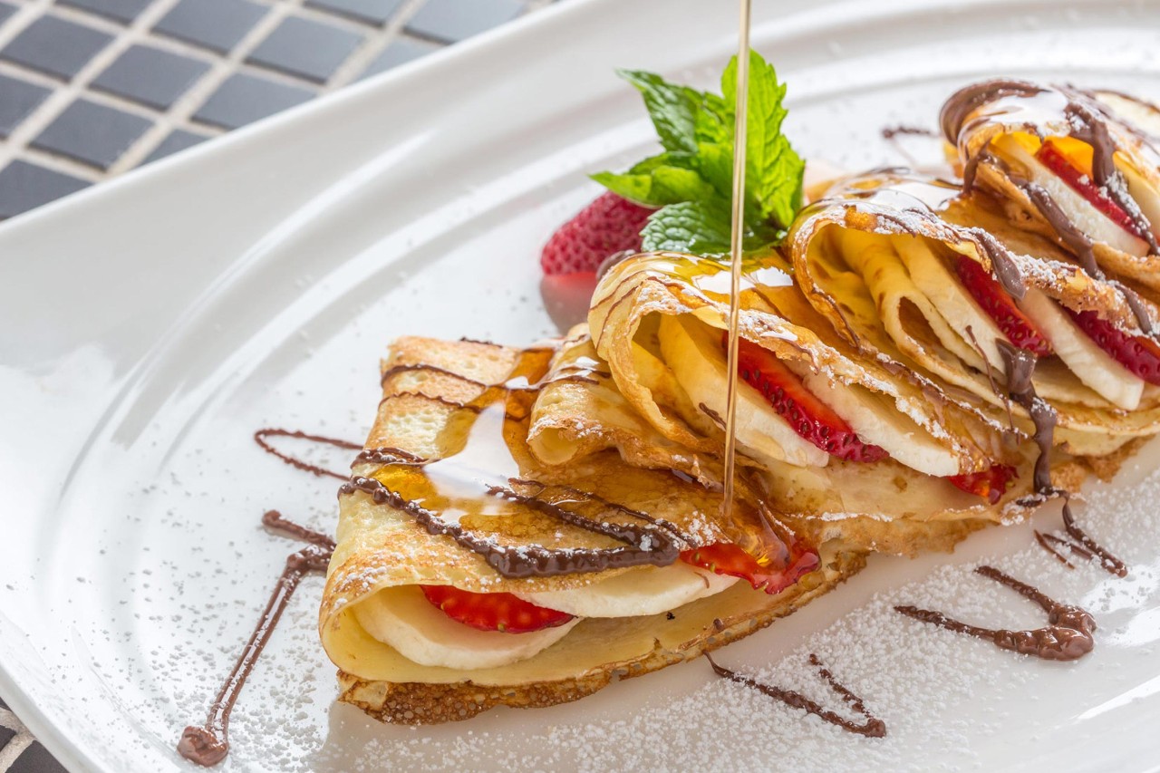 Close-up of folded crepes filled with strawberries and bananas, drizzled with syrup and garnished with mint on a white plate.