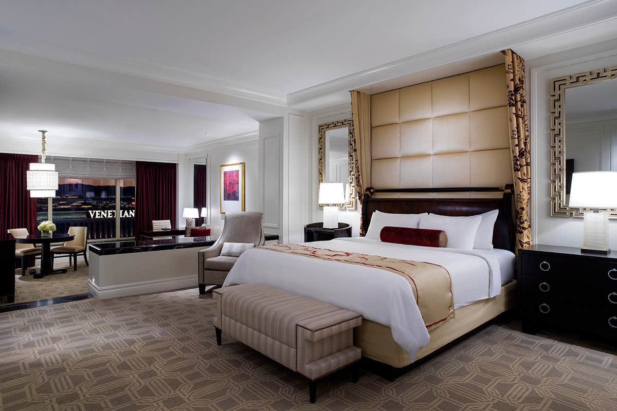 Luxurious hotel room with a king-sized bed, elegant decor, sitting area, and a view of the "Venetian" sign outside.