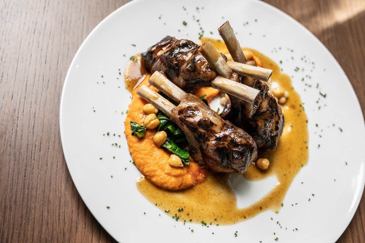 Grilled lamb chops on a white plate with chickpeas, spinach, and a bright orange puree, garnished with sauce and herbs.