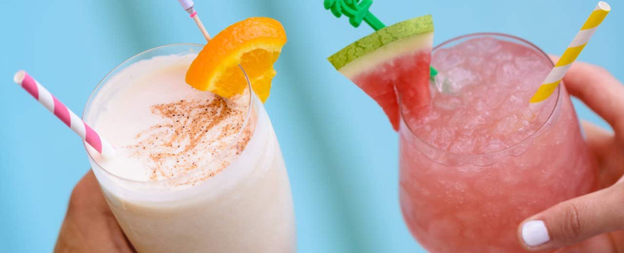 Two hands holding tropical drinks garnished with fruit slices against a blue background.