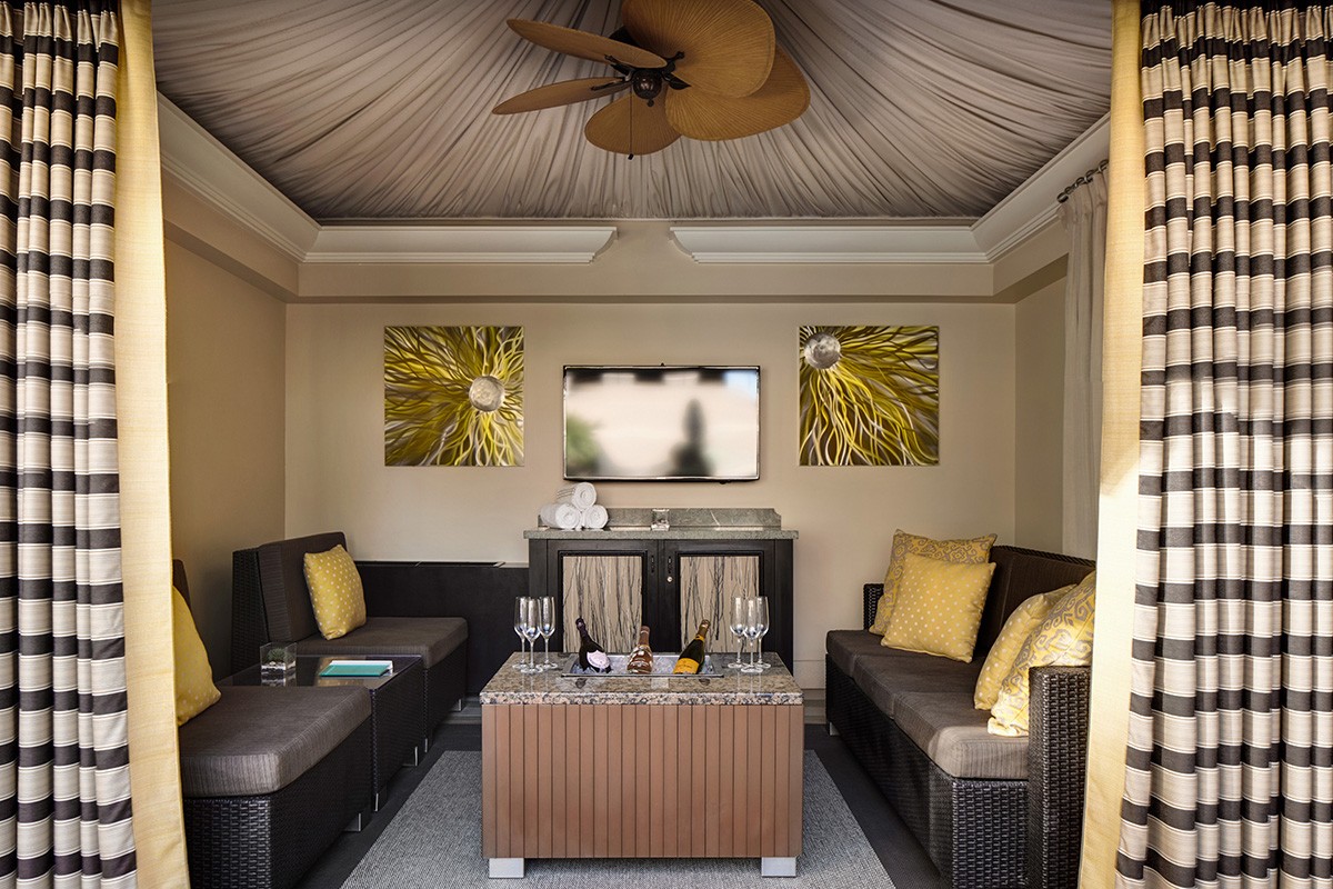 A cozy outdoor cabana with striped curtains, wicker furniture, yellow cushions, a TV, and a ceiling fan.