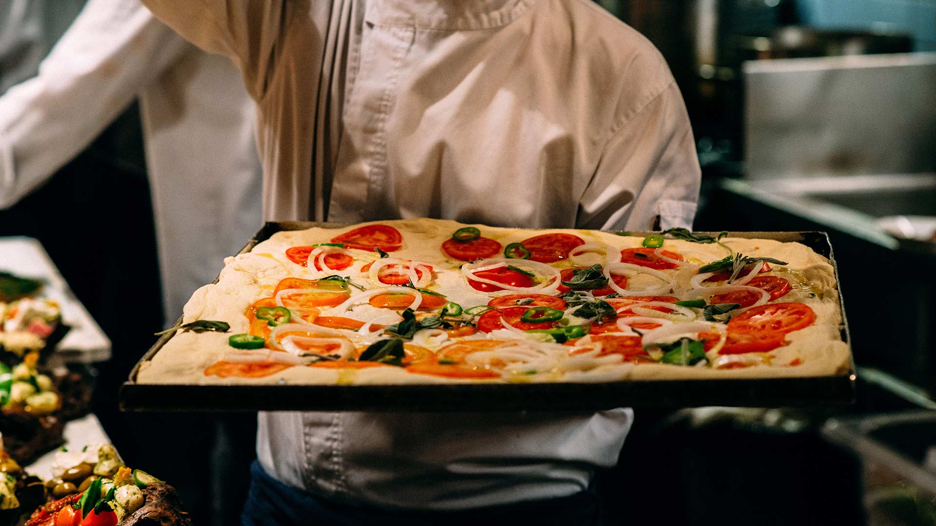 A chef holds a rectangular tray of uncooked pizza topped with tomatoes, onions, and green peppers.