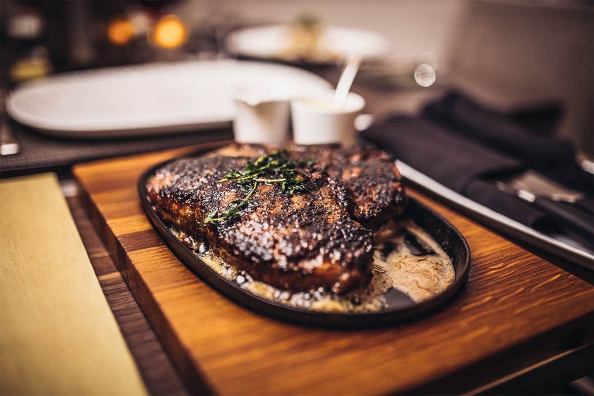 A sizzling steak garnished with herbs on a wooden board, accompanied by sauces and cutlery on a dining table.